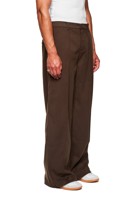 Männermodell trägt Exquisite Dress Pants Brown mit weitem Schnitt, Bundfalten, versetzter Seitennaht und feinem Reißverschluss, kombiniert mit weißen Sneakers, vor weißem Hintergrund.