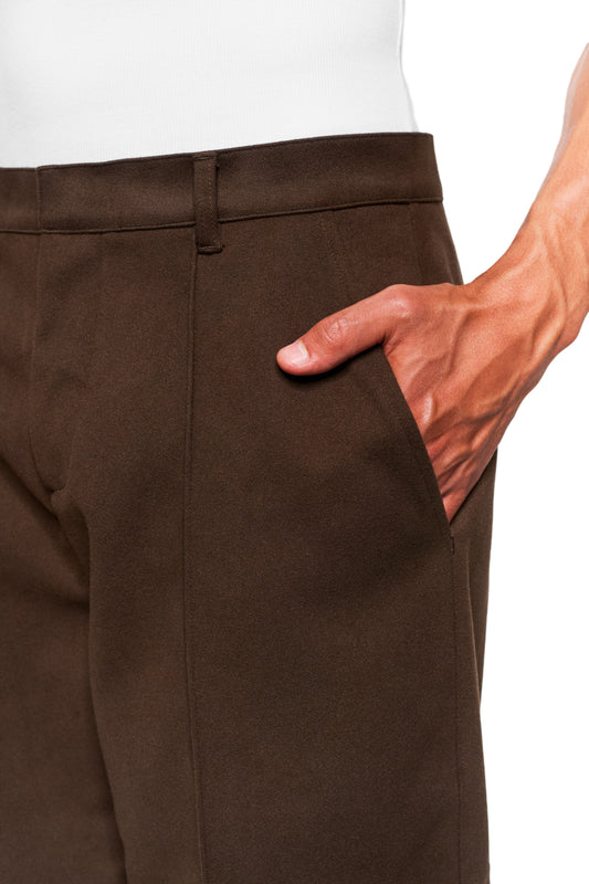 Person wearing Exquisite Dress Pants Brown with pleats and offset side seam. Right hand in trouser pocket, zipper at hem, embroidered OLAKALA icon visible.