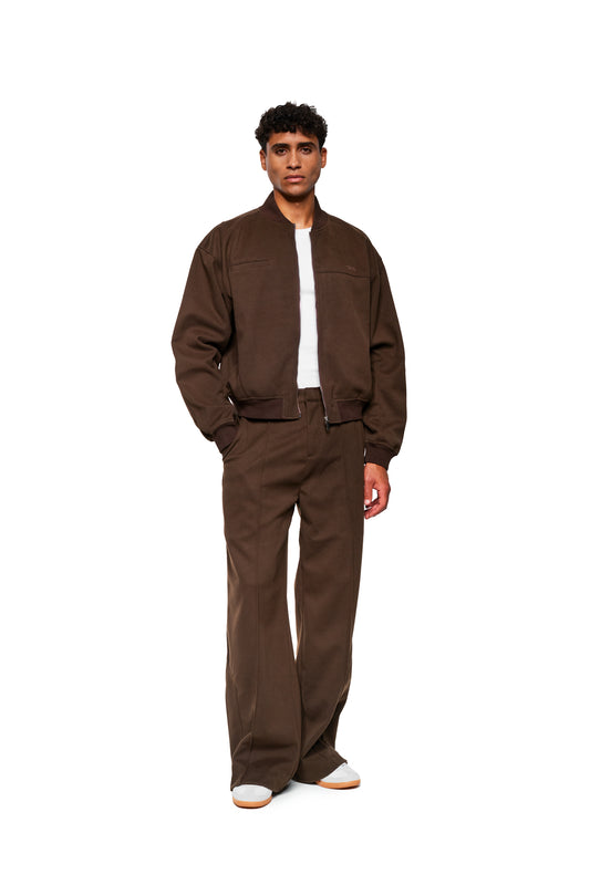 Men's model in Exquisite Bomber Jacket Brown with ribbed stand-up collar, chest pocket, zippered inner pocket and embroidered OLAKALA icon, combined with matching brown trousers.