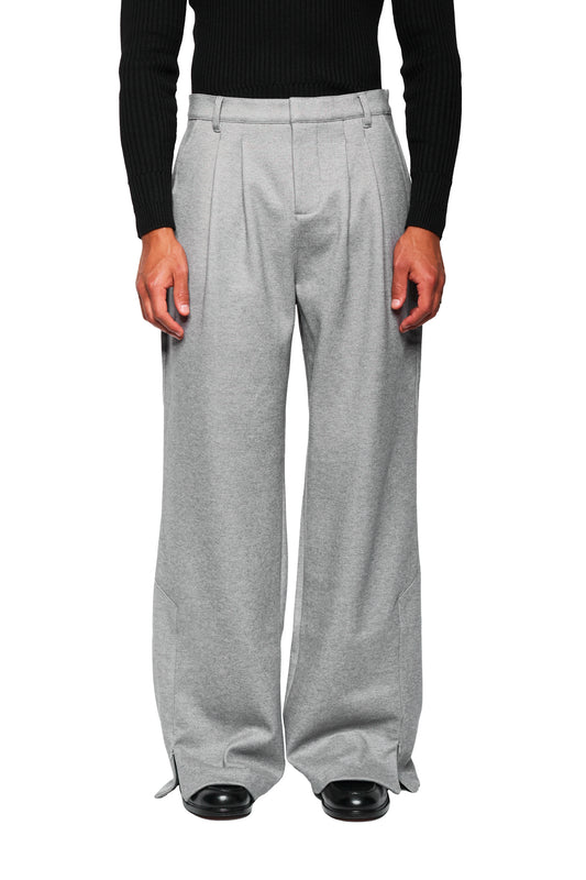 Exquisite Dress Pants Gray: Weite, graue Bundfaltenhose aus festem Köper, getragen mit schwarzem Pullover und Schuhen. Gesticktes OLAKALA-Icon an der Tasche sichtbar.