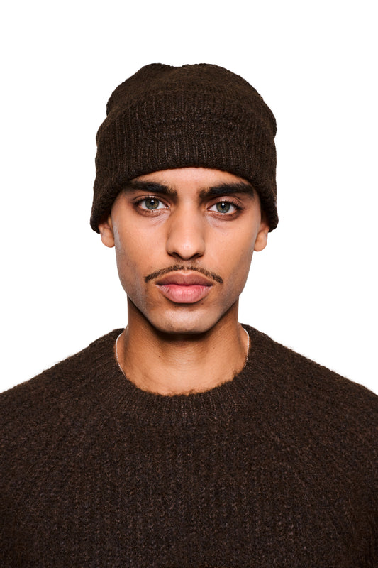Nahaufnahme eines jungen Erwachsenen mit dunkelbrauner Fuzzy Beanie Brown, die ein großes, geprägtes Logo zeigt, kombiniert mit passendem Wollpullover, im schlichten Design für kalte Tage.