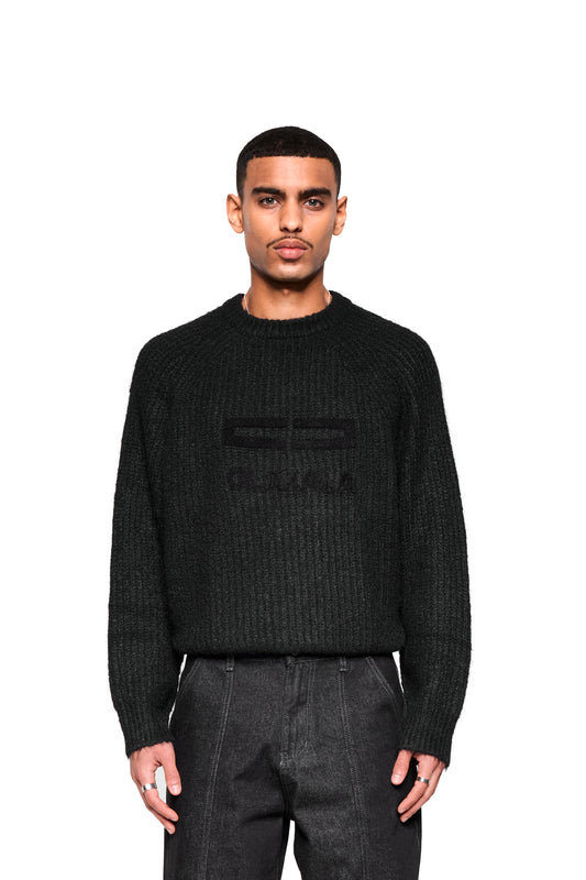 Junger Mann im Fuzzy Knit Sweater Black mit großer Logo-Stickerei, boxy Silhouette, leicht eingesteckt in dunkle Hose, trägt einfache silberne Ringe.
