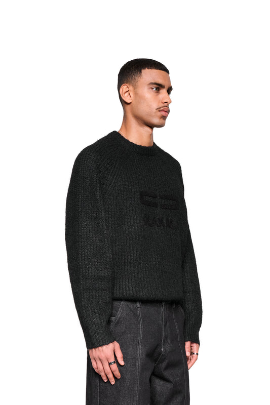 Junger Mann trägt Fuzzy Knit Sweater Black, ein kastig geschnittener, strukturierter Pullover mit großer Logo-Stickerei, kombiniert mit dunklen Jeans, präsentiert im OLAKALA-Stil.