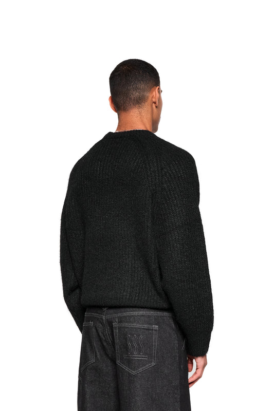 Rückansicht eines Mannes im Fuzzy Knit Sweater Black mit grobem Strick und cropped, boxy Silhouette, kombiniert mit Jeans und markanter Logo-Stickerei vorn.