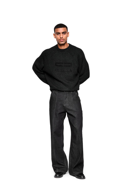 A man wears the Fuzzy Knit Sweater in black, featuring large embroidered logo details and a cropped, boxy silhouette, paired with high-waisted, wide-leg pants and black leather shoes.