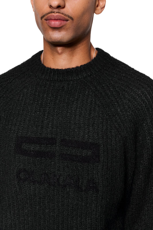 Person trägt Fuzzy Knit Sweater Black mit cropped, boxy Silhouette und großer, erkennbarer Logo-Stickerei „OLIMA“ auf der Vorderseite.