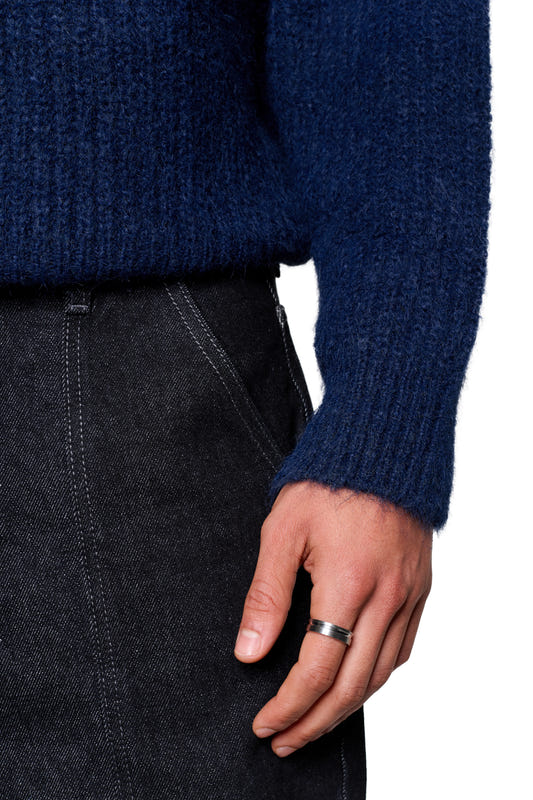 A person wearing a navy blue fuzzy knit sweater with a chunky texture and a modern, boxy silhouette is wearing a silver ring. The sweater is tucked into dark pants.