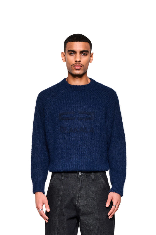 A male model wears a navy blue fuzzy knit sweater with a cropped, boxy silhouette and large embroidered logo, paired with high-waisted dark jeans.