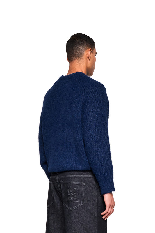 Rear view of a model wearing the Fuzzy Knit Sweater in Navy Blue, featuring a relaxed fit and textured surface, paired with dark jeans featuring an embroidered emblem.