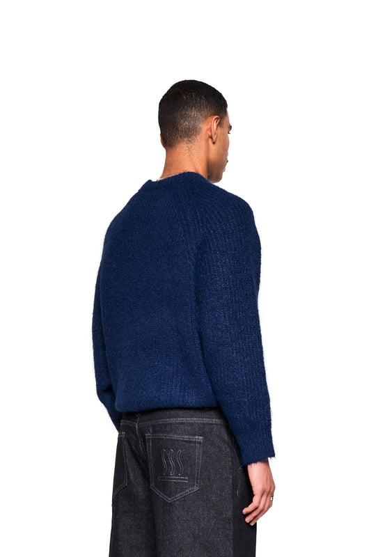 Rückansicht eines Modells im Fuzzy Knit Sweater Navy Blue, mit lockerer Passform und strukturierter Oberfläche, kombiniert mit dunklen Jeans mit einem gestickten Emblem.