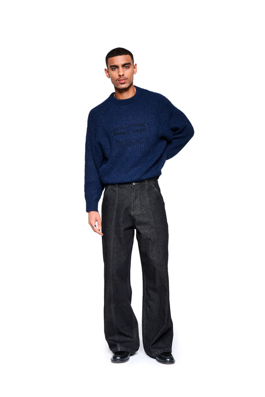 A male model wears the Fuzzy Knit Sweater in Navy Blue with large embroidered logo, paired with black jeans and leather shoes.