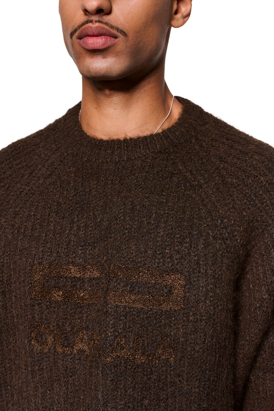 Mann in einem Fuzzy Knit Sweater Brown mit großem Logo-Stick auf der Brust und silberner Kette um den Hals.