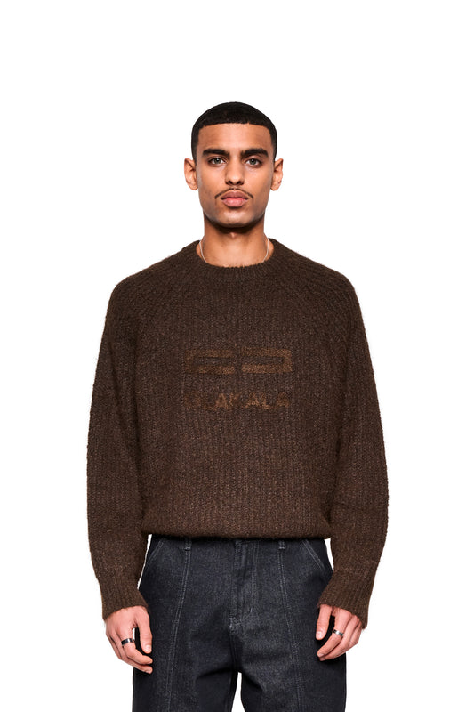 Junger Mann trägt Fuzzy Knit Sweater Brown mit lockerer Passform und großer Logo-Stickerei, kombiniert mit dunkler Jeans, vor weißem Hintergrund.