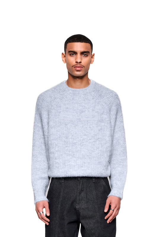 Junger Mann trägt den Fuzzy Knit Sweater Gray mit cropped, boxy Silhouette und großer Logo-Stickerei. Perfekte Verbindung von Komfort und modischem Statement.