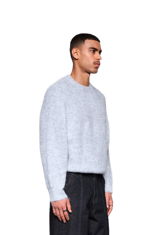 Junger Mann im Profil trägt den Fuzzy Knit Sweater Gray mit cropped, boxy Silhouette und großer Logo-Stickerei, kombiniert mit dunklen Hosen und Ringen.