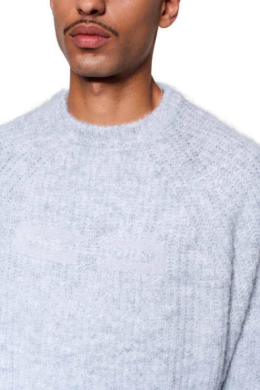 Person trägt den Fuzzy Knit Sweater Gray mit cropped, boxy Silhouette und weicher Struktur. Auffällig ist die große Logo-Stickerei vorne.