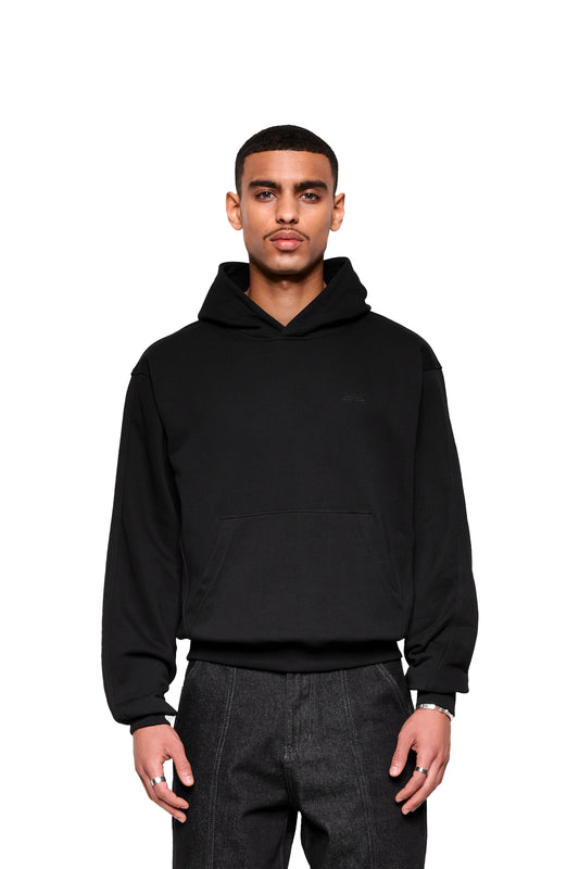 Junger Mann im Mokka Hoodie mit Kängurutasche und optimierten Nähten für Komfort, trägt schlichte Ringe und Armband, stehend mit entspannten Armen.