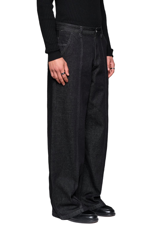 Person trägt Raw Denim Pants Black mit weitem Bein und mittlerer Leibhöhe. Auffällige Frontnähte und French-Pockets sichtbar. Jeans über schwarzen Lederschuhen drapiert.