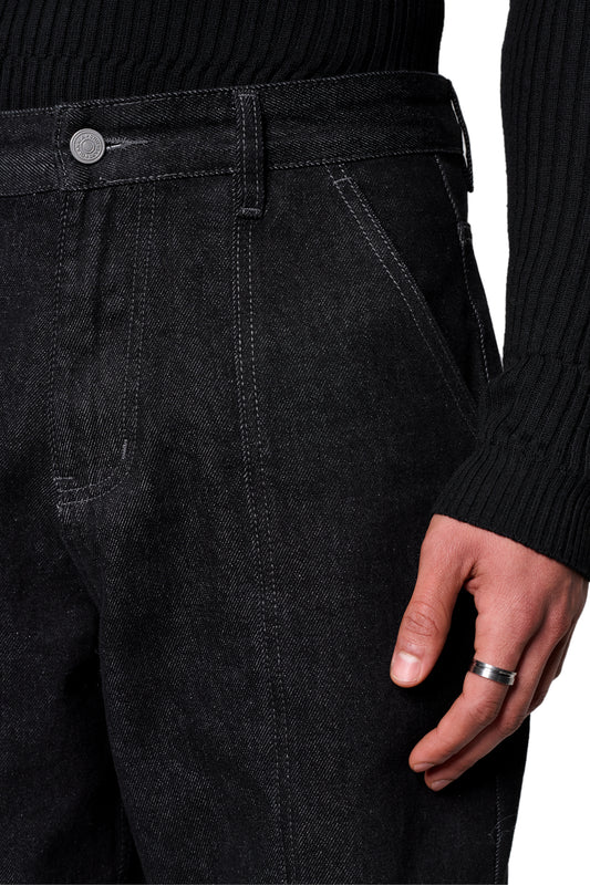 Nahaufnahme der Raw Denim Pants Black mit sichtbaren Gürtelschlaufen, Vordertaschen, silbernem Knopf und kontrastierenden Nähten, getragen zu einem schwarzen gerippten Pullover.