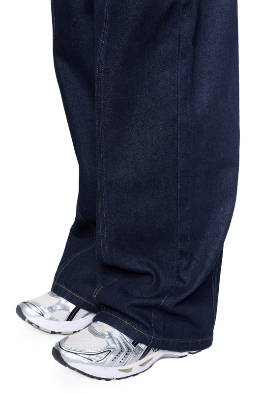 Beine in Raw Denim Pants Indigo mit lockerer Passform, geradem Bein über silber-weißen Sneakern, betonen markante Nahtdetails und French-Pockets.