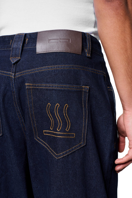Rückansicht der Raw Denim Pants Indigo mit goldener Stickerei auf der Gesäßtasche und braunem Leder-Patch am Bund, getragen mit weißem Tanktop.