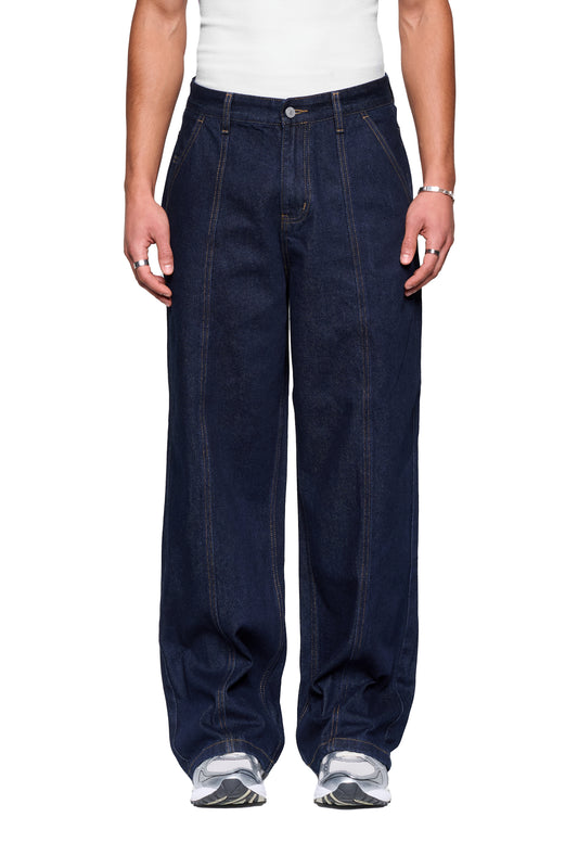 Person trägt Raw Denim Pants Indigo: weite, dunkle Jeans mit markanten Nähten, French-Pockets, und dezentem Stickdetail, kombiniert mit weißem Tanktop und grauen Sneakers.