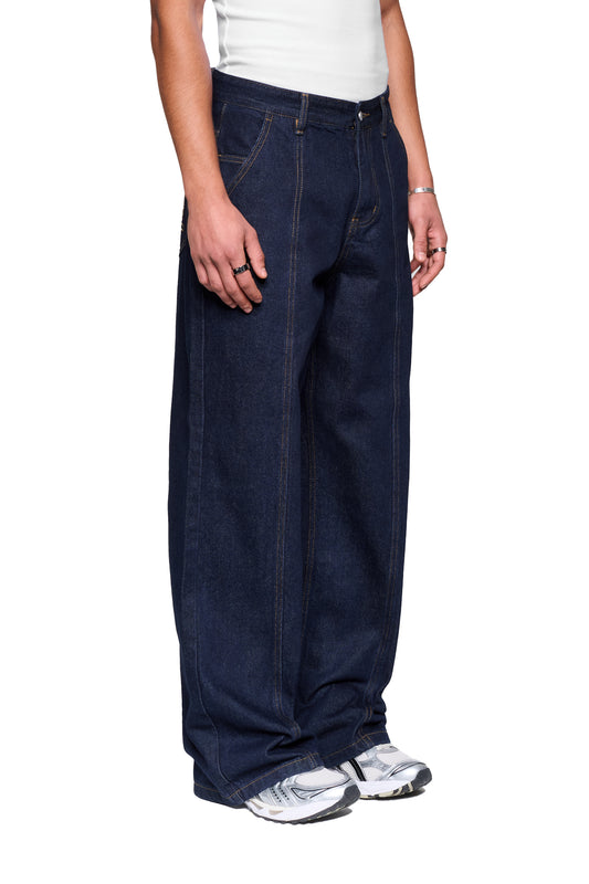 Person trägt Raw Denim Pants Indigo mit extrem weiten Beinen und sichtbaren Nähten, kombiniert mit weißen Sneakers, silbernen Ringen und einem schmalen Armband.