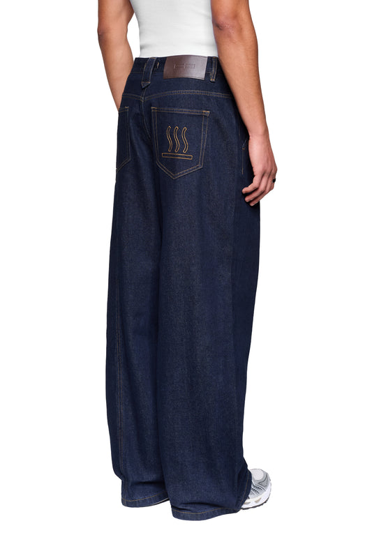 Person in Raw Denim Pants Indigo mit weiten Beinen, gelben Nähten und Stickerei auf der Gesäßtasche, getragen mit grauen Sneakers.