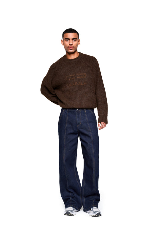 Männermodell trägt Raw Denim Pants Indigo mit weitem Bein, französischen Taschen und markanten Nähten, kombiniert mit einem dunkelbraunen Strickpullover und weißen Sneakers.