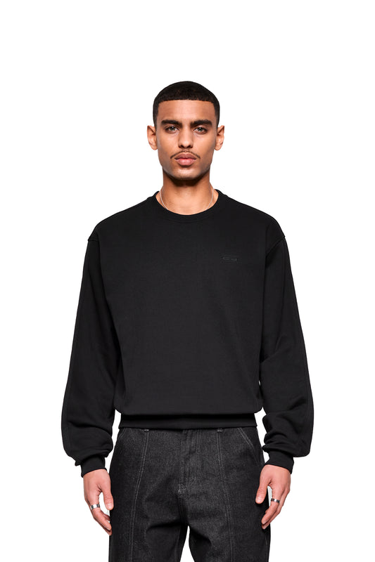 Junger Mann trägt den Mokka Sweater, einen schwarzen Crewneck-Pullover von OLAKALA, kombiniert mit dunklen Hosen, silberner Kette und Ringen.