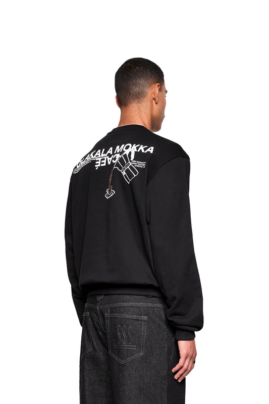 Rückansicht des Mokka Sweaters mit MOKKA CAFÉ Grafik und Moka-Kanne. Kombiniert mit dunklen Jeans, die ein wellenförmiges Stichmuster aufweisen.