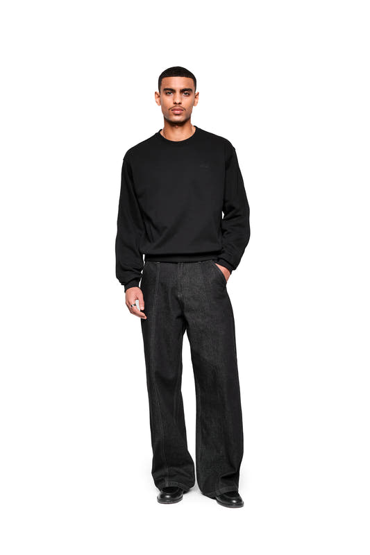 A male model wears the Mokka Sweater, a black long-sleeve sweater with special seams for comfort, paired with wide-leg pants.