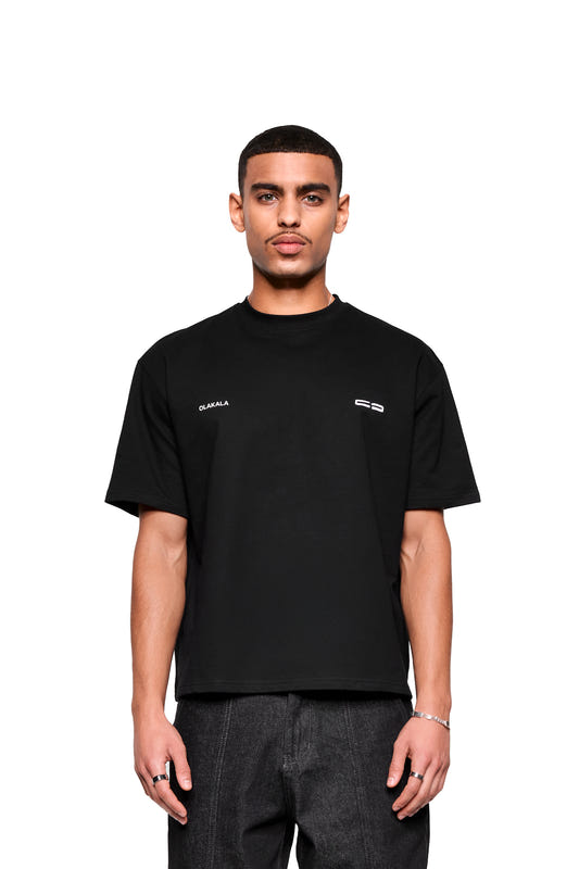 Junger Mann im Mokka T-Shirt Black von OLAKALA, mit kleinem Logo-Print vorne, trägt dunkle Jeans, Silberarmband und Ringe, schaut neutral in die Kamera.