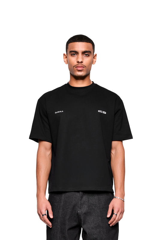 Junger Mann im Mokka T-Shirt Black von OLAKALA, mit kleinem Logo-Print vorne, trägt dunkle Jeans, Silberarmband und Ringe, schaut neutral in die Kamera.