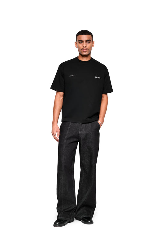 Mokka T-Shirt Black im boxy Schnitt mit OLAKALA-Logo, kombiniert mit weiten Jeans und Loafers. Modell trägt Ring und Armband.