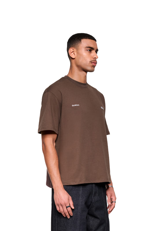 Junger Mann im Profil trägt das OLAKALA Mokka T-Shirt Brown mit lockerem Fit und dezentem Logo, kombiniert mit dunklen Jeans und schlichten Silberringen.