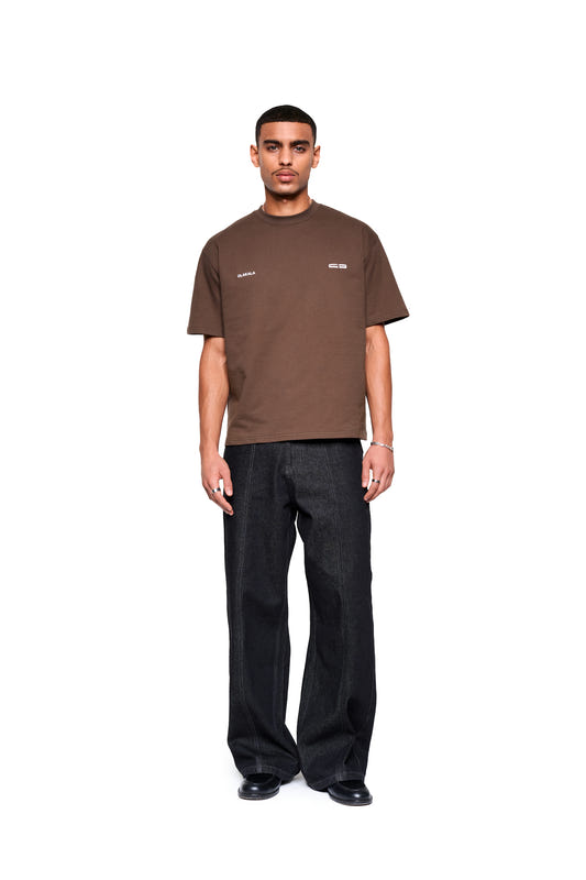 Mann trägt Mokka T-Shirt Brown mit boxy Fit und dezentem Logo-Print vorne, kombiniert mit dunkelblauen Jeans und schwarzen Schuhen, präsentiert von OLAKALA.