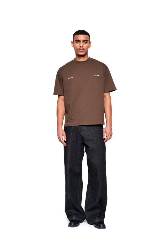 Mann trägt Mokka T-Shirt Brown mit boxy Fit und dezentem Logo-Print vorne, kombiniert mit dunkelblauen Jeans und schwarzen Schuhen, präsentiert von OLAKALA.