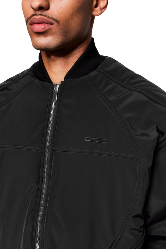 Mann trägt Void Bomber Jacket Black mit Reißverschluss, geripptem Kragen und dynamischen Nähten, die dreidimensionale Form betonen. Ideal als Statement-Piece oder Basics-Ergänzung.