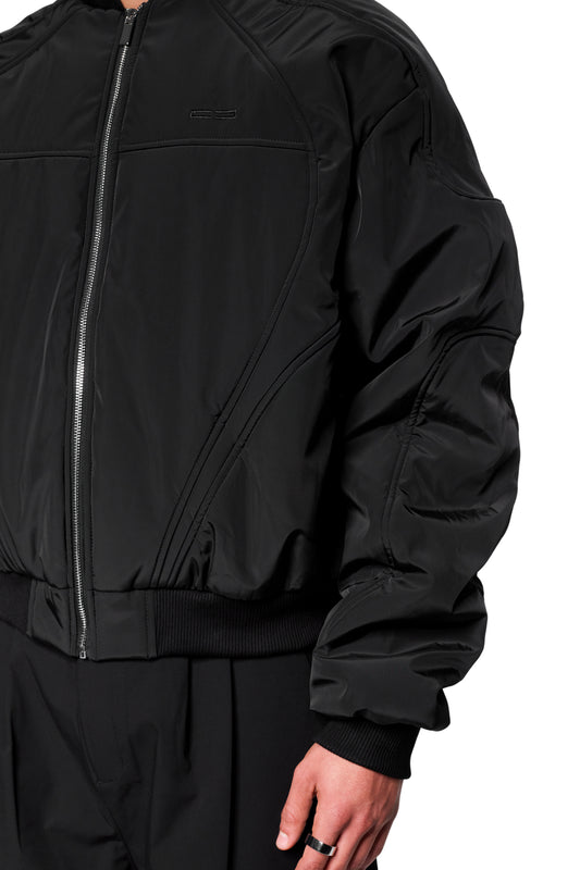 Person trägt die Void Bomber Jacket Black mit Reißverschluss, gerippten Bündchen und dezenten Nähten, die der Jacke eine dynamische, dreidimensionale Form verleihen.