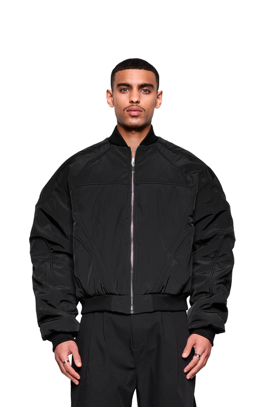 Junger Mann trägt die Void Bomber Jacket Black mit Reißverschluss, gerippten Bündchen und dynamischen Nähten, kombiniert mit silbernen Ringen und passenden Hosen.