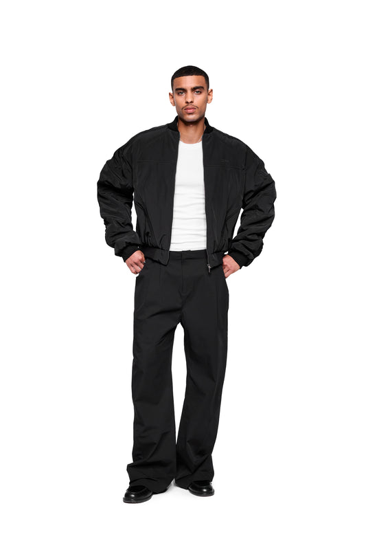 Modell trägt Void Bomber Jacket Black mit dynamischen Teilungsnähten, geripptem Kragen und elastischen Bündchen, kombiniert mit weißem T-Shirt und locker geschnittenen schwarzen Hosen.