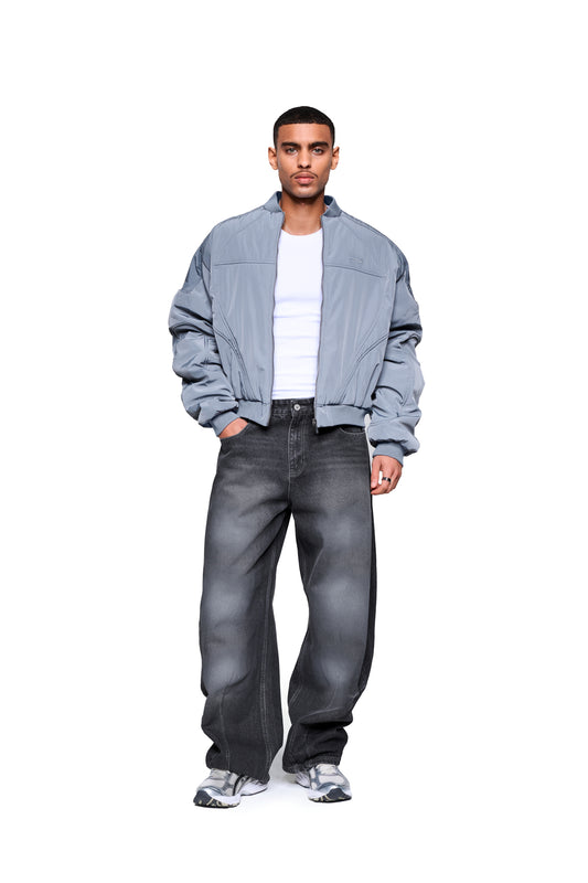 Junger Mann trägt die Void Bomber Jacket Gray mit dynamischen Nähten und Rippbündchen, Hände in Taschen, steht vor weißem Hintergrund, OLAKALA® Stil.