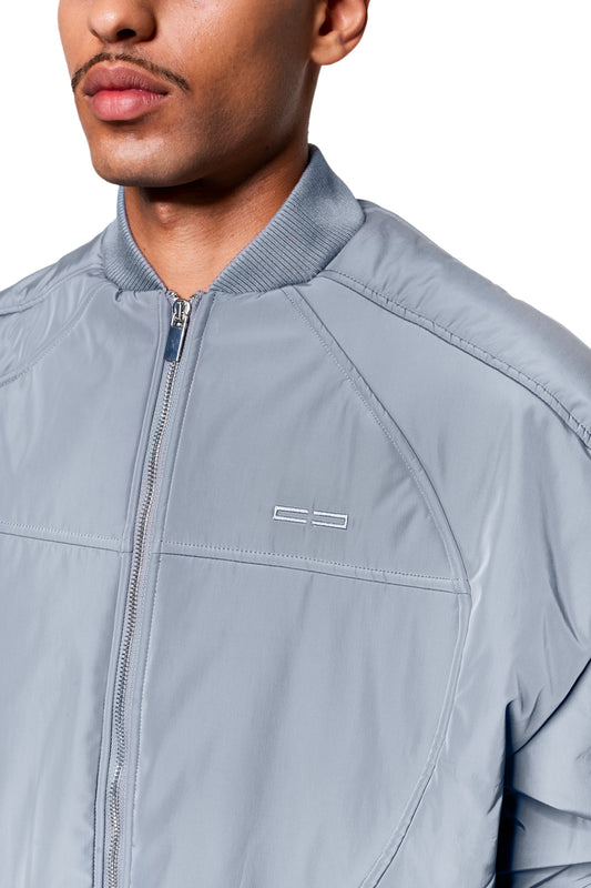 Person trägt Void Bomber Jacket Gray mit Rippenkragen, Metallreißverschluss und dezentem Logo links auf der Brust, betont durch dynamische Teilungsnähte.