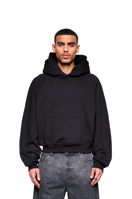 Junger Mann trägt OLAKALA Double Hood Hoodie Black mit Kängurutasche und Raglanärmeln, locker geschnitten, vor weißem Hintergrund stehend.