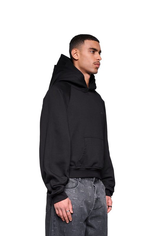 Junger Mann im Profil trägt den Double Hood Hoodie Black mit Kängurutasche und asymmetrischem Bund von OLAKALA, kombiniert mit dunklen Jeans.