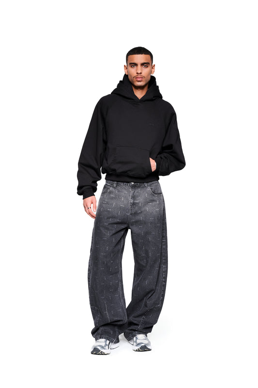 Mann trägt OLAKALA Double Hood Hoodie Black mit Kängurutasche und Raglanärmeln, kombiniert mit locker sitzenden Jeans und Sneakers, vor weißem Hintergrund.