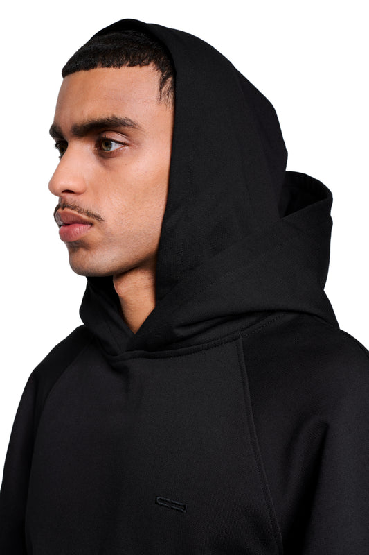 Junger Mann im Profil trägt schwarzen Double Hood Hoodie mit OLAKALA-Stickerei auf der Brust, Kängurutasche und asymmetrischem Bund.