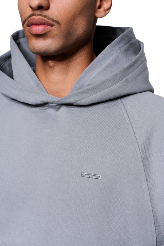 Person trägt Double Hood Hoodie Gray mit OLAKALA-Logo auf der Brust, Raglan-Ärmeln, Kängurutasche und umgelegter Kapuze. Unisex, weit und kurz geschnitten.