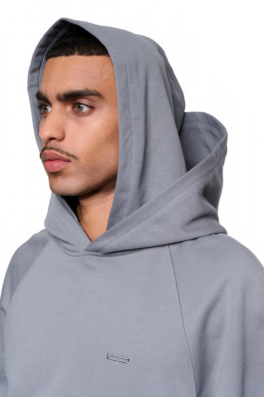 Junger Mann im Double Hood Hoodie Gray mit Kapuze, OLAKALA-Logo auf der Brust. Lockerer Schnitt mit Kängurutasche und Raglanärmeln.